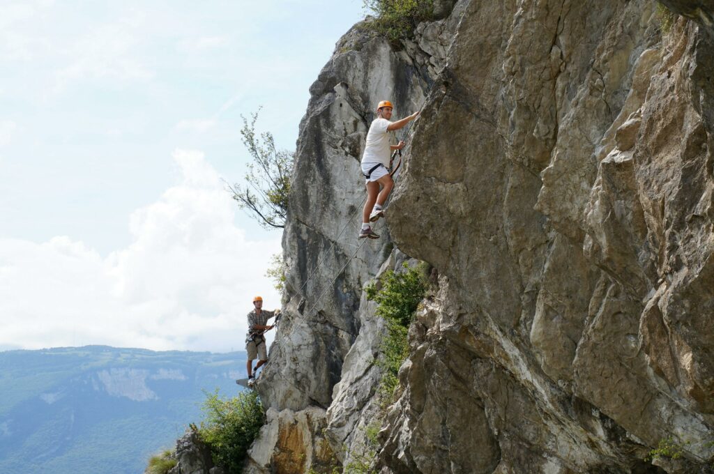 Activités Two men climbing a steep mountain rock face with safety gear under a bright summer sky.